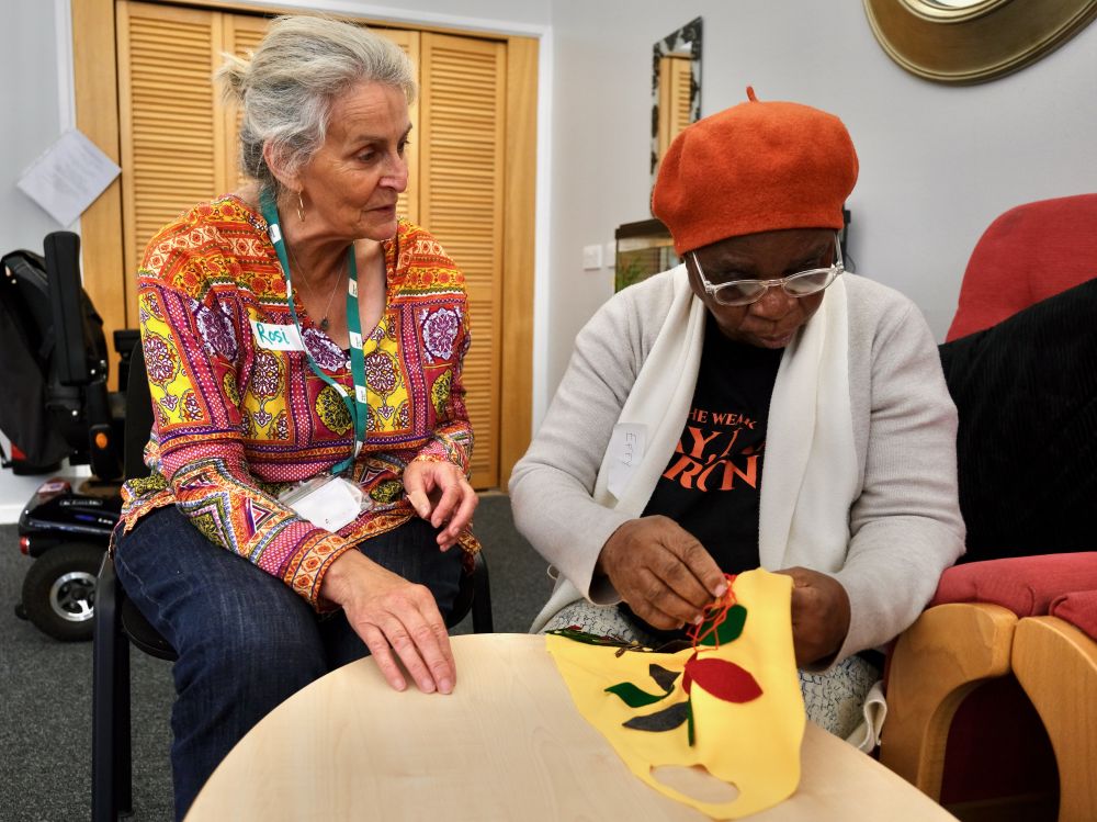 A silver haired facilitator helps a woman stitching a quilt patch
