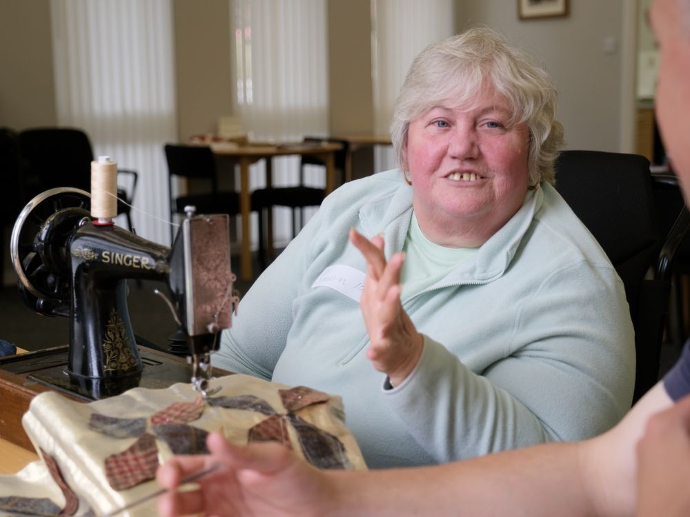 A woman in a wheelchair gestures her hand forward. There is an old sewing machine in the foreground